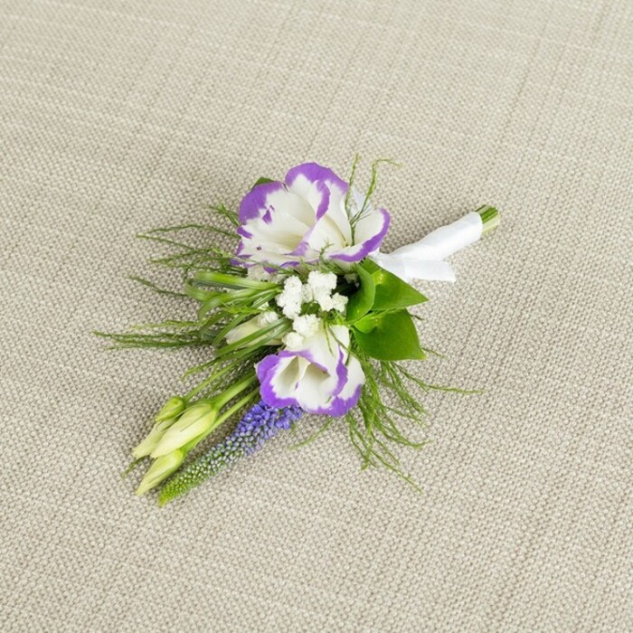 A finely detailed boutonniere is set against a pale beige, gently textured fabric that looks like natural linen, its understated background echoing the relaxed elegance of a St Mary Cray wedding gathering. At the centre of this small floral piece sit two lisianthus blooms, fully opened to reveal layered, rose-like petals. Their bases are pure white, silky and luminous, while each ruffled petal tip is edged with a rich band of deep purple, creating a vivid halo that feels both romantic and contemporary. Around these focal flowers, clusters of tiny, pristine white filler blossoms-similar to baby's breath-fill the gaps between petals, contributing a soft, cloud-like texture and a classic bridal touch. To one side, a slim vertical stem of grape-hyacinth-style flowers rises, its tiny bell-shaped florets packed tightly together in a column of deep lavender-purple that adds depth and visual rhythm. Fine, feathery greenery, likely asparagus fern or a similar foliage, fans around the blooms in airy sprays, lending movement and a natural, garden-picked charm, as though the stems were gathered from an early-spring border near the River Cray. Two pale green buds, still closed, are tucked into the design, symbolising freshness and the promise of blossoms yet to open as the day unfolds. The stems are aligned neatly and wrapped with a band of crisp white satin ribbon, the smooth fabric catching the light and ending in a small, precise bow that signals expert florist craftsmanship. Prepared to be pinned to a lapel, this boutonniere feels ideal for a groom, groomsman, or formal guest in St Mary Cray seeking something understated, refined, and quietly sentimental, perfectly captured for use on a local florist website or wedding inspiration guide.