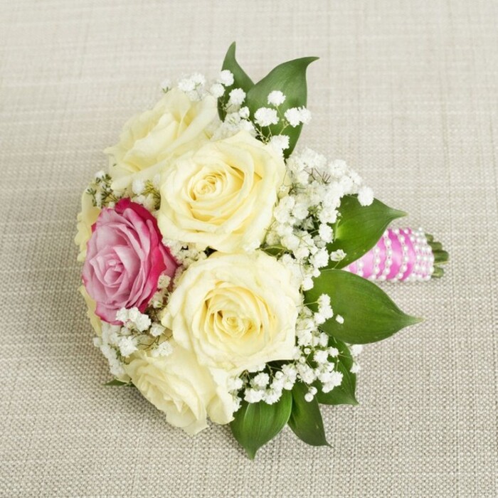 Viewed from above, this meticulously crafted bridesmaid bouquet rests on a soft beige linen evocative of St Mary Cray's welcoming community halls. The centrepiece features six creamy, unopened roses, their petals unfurling with an air of newness and promise-each one lovingly arranged by local florists. Slightly off-centre sits a vibrant fuchsia-pink rose, its bold colour immediately captivating yet perfectly at harmony with the gentler cream blooms. Sprigs of airy white baby's breath weave generously throughout, imparting an ethereal quality that softens the fuller roses and recalls meadows near Orpington. Glossy, deep green leaves form a verdant frame, offering natural structure and elegance. The bouquet's handle is wrapped with a plush fuchsia-pink satin ribbon that catches the light, spiraled perfectly with iridescent white pearls for a note of traditional charm. Set against the subtle fabric, the bouquet emanates warmth, happiness, and refined style-ideal for sophisticated weddings or celebrations in St Mary Cray. The thoughtfully chosen colours and textures make it a fresh and romantic piece certain to inspire local couples planning their unforgettable day.