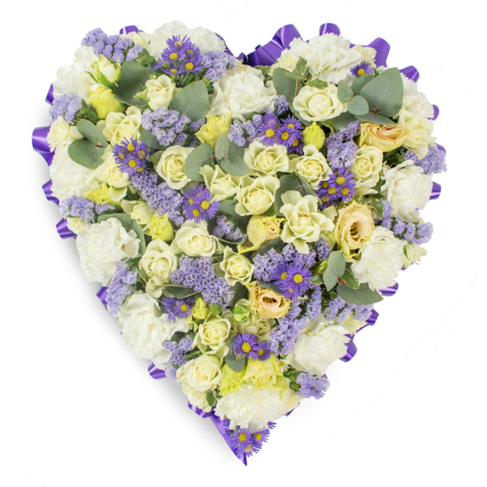 Against a spotless white studio field, this heart-shaped sympathy spray is densely composed and precisely finished, its outer edge defined by a continuous band of deep purple satin ribbon that forms a refined, graceful border. Inside that band, the composition relies on a soft, reassuring palette: predominantly cream and pale yellow roses arranged close together to form a plush, layered surface, their petals ranging from tightly held buds to softly open blooms that suggest tenderness and the passage of time. Interspersed throughout are full white carnations, their ruffled texture adding traditional volume and a familiar, comforting silhouette. Complementing the lighter tones are plentiful clusters of lavender-coloured statice, whose tiny, papery blooms create airy texture and diffuse lilac notes across the heart, alongside small purple daisy-like flowers with vivid yellow centres that provide rhythmic, deeper colour accents. Blue-green eucalyptus leaves are artfully placed to punctuate the floral mass, their rounded shapes and silvery sheen offering contrast and a hint of calming scent. The arrangement sits under gentle, diffuse lighting that highlights petal structure and the satin ribbon's soft reflection without dramatic shadows, reinforcing a mood of peaceful remembrance. Crafted with measured attention to balance, colour, and form, this floral heart would be appropriate for funeral services, memorial tables, or graveside placement - an emblem of respect and lasting affection from local makers who understand the quiet needs of families in St Mary Cray and the surrounding community.