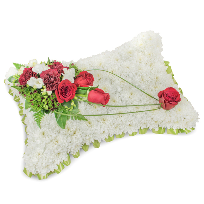 A dignified memorial cushion, shaped as a soft rectangle and photographed on a crisp white backdrop, is formed from hundreds of immaculate white chrysanthemums packed so closely the surface reads as plush and cloud-like. Along the gently rounded rim the hint of a pale green satin ribbon or trimmed foliage provides a subtle frame, delicate yet deliberate. Anchoring the composition in the top left, a luminous cluster of deep red roses and dark burgundy carnations creates a focal point: the roses' velvety petals and the carnations' layered frills offer a lush contrast to the snowy chrysanthemum base. Small sprays of white freesia or tiny spray chrysanthemums nestle among the red blooms, their tiny cups offering brightness and a whisper of scent. Behind and between these flowers, verdant greenery-feathery fern fronds, glossy salal or eucalyptus leaves-fans outward to support and set the colours free. From the clustered spray a graceful band of bright green bear-grass or decorative stems crosses diagonally toward the pillow's lower right, forming an elegant sweep that culminates in a single, perfectly formed red rose resting at the corner. The arrangement conveys calm, respect and personal tenderness, intended for a funeral service or chapel tribute; it suggests a quiet morning in St Mary Cray near the River Cray, where such memorial cushions are often placed to honour a life with both simplicity and care.
