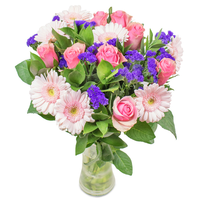 An exquisite hand-arranged bouquet from a trusted St Mary Cray florist, this display stands proudly in a clear glass vase set before a crisp, white backdrop. The centerpiece features approximately seven to nine medium and pastel pink roses, their petals still partly furled, showing off the signature spiral form that speaks to the time-honored beauty of traditional gardens found near Poverest Road. Embracing the roses are five to seven pale pink gerbera daisies, notable for their expansive, rounded heads and radiating petals, with nearly white centers that brighten the overall composition. Among the main blooms, clusters of brilliant purple statice deliver a striking texture and depth, providing vibrant punctuation to the gentler tones. Dense, deep green foliage fills the base, framing the flowers with an abundant, fresh burst of greenery that conjures the wild charm of local riverside walks. The vase, crystal clear and filled with water, completes a sense of lively freshness-suggesting a recent delivery, ready to grace any home or office near St Mary Cray. Ideal for birthdays, anniversaries, or Mother's Day, this arrangement radiates joy, celebration, and the enduring warmth of heartfelt affection.