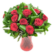 A lush, rounded bouquet of more than a dozen deep crimson roses sits proudly in a tall, slender ruby-red glass vase, each bloom opened to a perfect, velvety cup. The petals are rich and wine-dark at the heart, softening to satin edges that catch the even, diffused studio light and reveal delicate veining and layered texture. Between the rose heads an abundance of glossy dark green foliage creates a structured, verdant framework: narrow, flat leaves like Ruscus or Salal form a dense backdrop while broader, emerald Aspidistra-style leaves are skillfully curled and spiralled into architectural loops that cradle and highlight individual blooms. These folded leaves introduce a modern sculptural rhythm against the classic romance of the roses, their glossy surfaces reflecting subtle highlights from the vase's ribbed, twisted glass. The vase itself is translucent ruby red with vertical swirls that refract light and deepen the bouquet's passionate palette; its narrow waist and flared top give an elegant silhouette that stabilises the composition. Set against a clean white background, the contrast between the deep crimson and lush greens is striking and immediate-every petal edge and folded leaf stands out with clarity. As a local florist might say, this arrangement reads as a quiet, sophisticated declaration of love: the scent suggested is soft and classic, the overall feeling intimate and ceremonial. Picture this as an anniversary gift collected near St Mary Cray station or left on a bedside table after a walk along the River Cray-timeless, luxurious, and carefully composed for someone who loves refined romance.