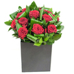 I describe this arrangement as a study in confident simplicity: a tight cluster of crimson roses, each bloom unfolding in velvety spirals, sits as the unmistakable heart of the design. Around them, broad glossy green leaves have been carefully curled and overlapped, creating waves of vibrant emerald that both frame and push the eye toward the rich red centers. The flowers are presented in a matte black, square box vase that gives the composition a grounded, modern silhouette - its flat planes and soft finish contrasting with the lush sheen of petals and foliage. The studio lighting here is crisp and contemporary, catching tiny highlights on the leaf surfaces and letting the crimson tones glow with an almost velour depth. I often think of placing this on a low table in a St Mary Cray living room after a walk along the River Cray: the scent you'd notice is the familiar, classic perfume of roses softened by the green, almost tropical lift of the leaves. Running fingers near the blooms, you'd sense the velvet softness of each petal against the firmer, slightly waxy texture of the greenery. As a florist I arranged each leaf to create movement and balance - the result reads as romantic and composed, ideal for an anniversary, a heartfelt apology, or to bring a deliberate note of warmth to a modern office near the High Street. The bouquet's restraint and the tactile contrast between soft petals and glossy foliage make it a memorable, intimate statement without ostentation.